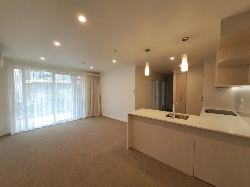 Apartment kitchen and open plan lounge with timber toned cabinetry, breakfast bar and balcony doors